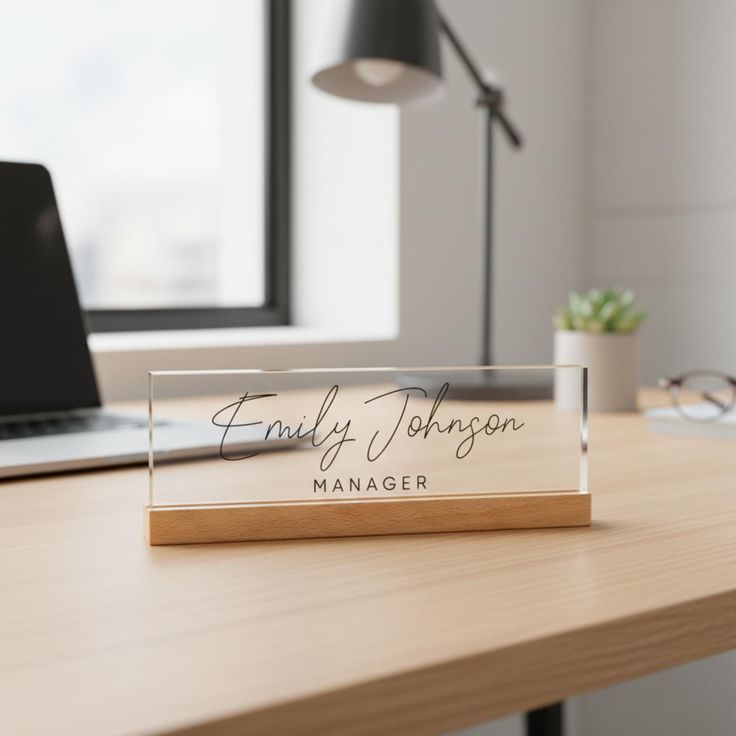 A minimalist acrylic desk name plate with clear, clean lines and black engraved lettering, placed on a tidy workspace