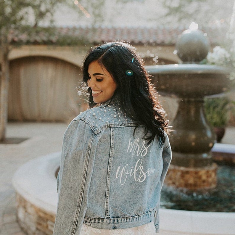 May include: A light blue denim jacket with pearl embellishments on the shoulders. The back of the jacket has white lettering that says 'Mrs. Wilson'.