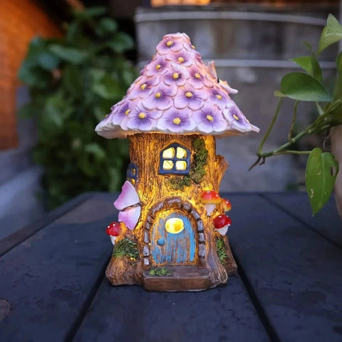 A small, illuminated fairy house with a purple flower-covered roof sits on a dark surface at night. It has a glowing blue door and window, and decorative mushroom and butterfly accents.