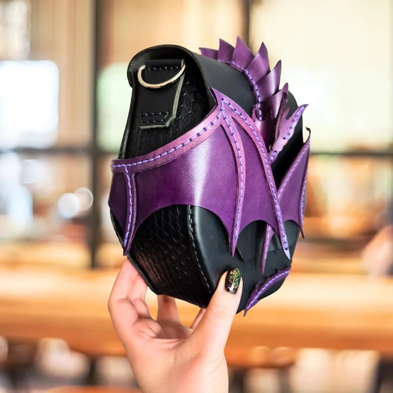 May include: A black leather crossbody bag with purple dragon wing and spine accents. The bag features a flap closure with a gold-colored clasp. The dragon wings are stitched onto the front, and the spine runs along the top edge. The bag is held by a hand with a glittery nail polish.