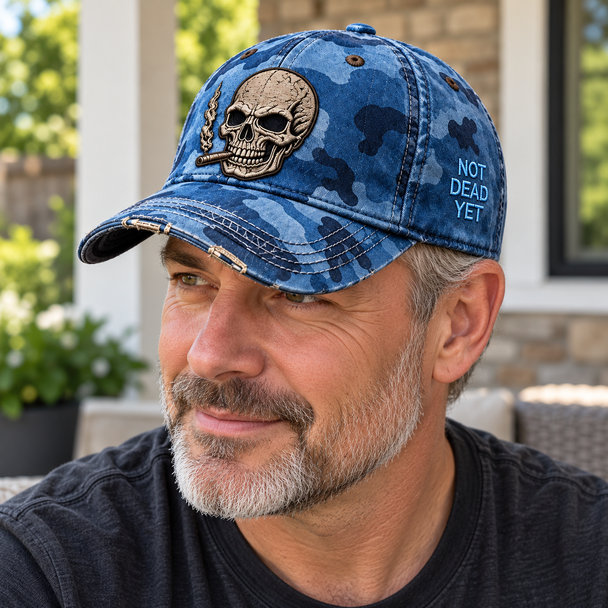 A mature man wearing the blue camouflage Not Dead Yet cap outdoors with the front skull and side lettering visible