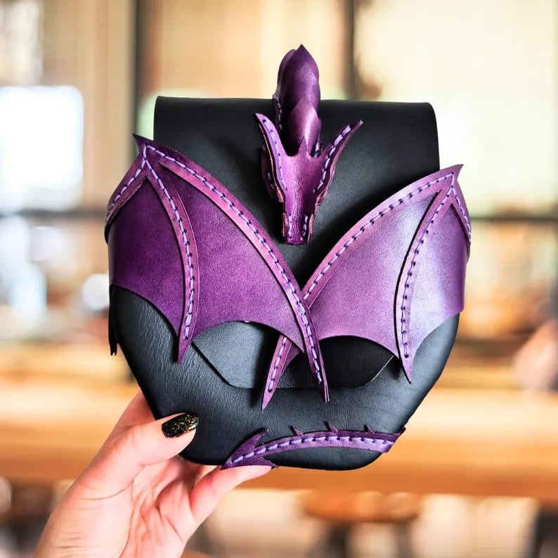 May include: Black leather pouch with purple dragon wing and head accents. The pouch has a flap closure and is decorated with stitched details. The dragon wings are on the front, and the head is on the top. The wings and head are a vibrant purple color, contrasting with the black pouch.