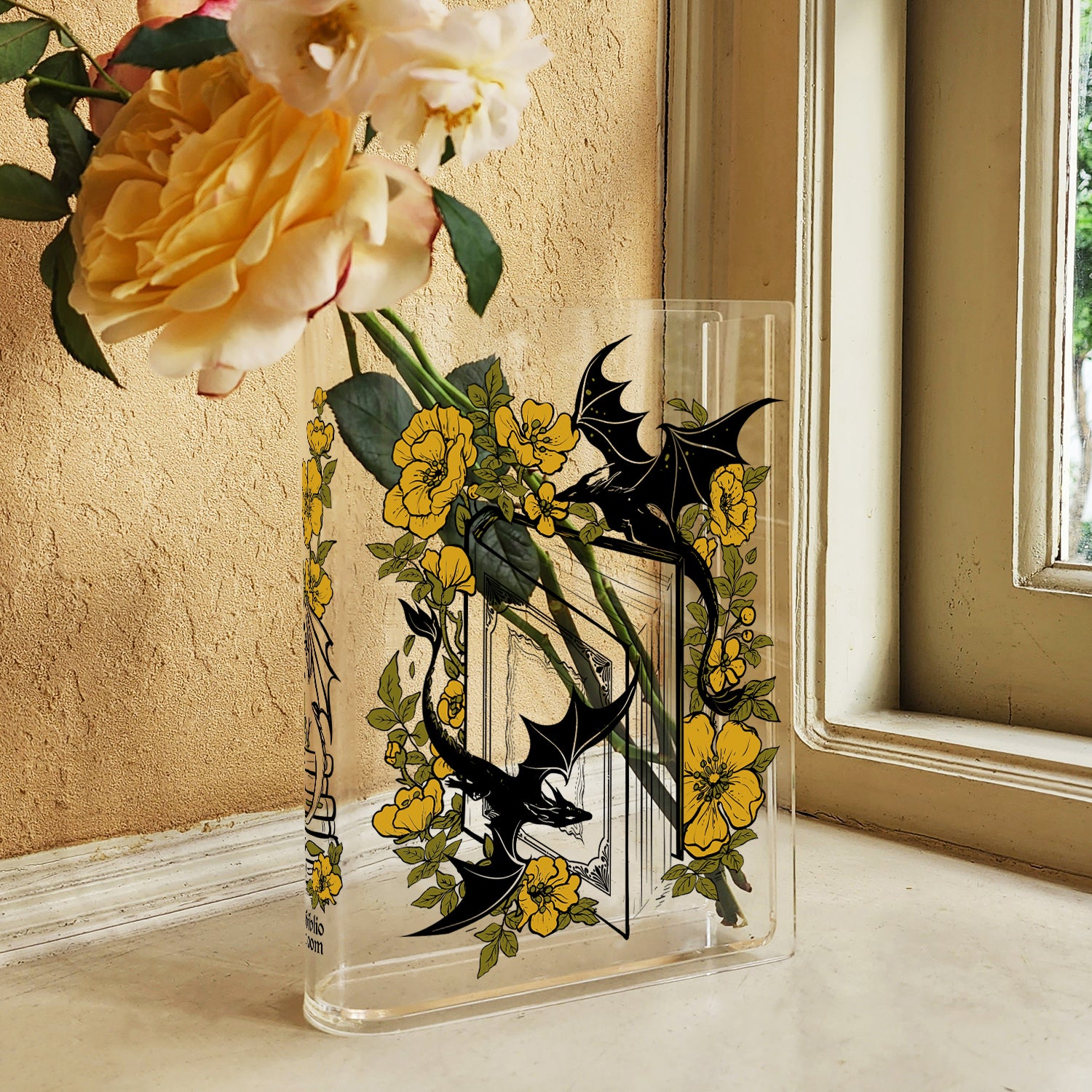 Acrylic book-shaped vase with dragon and yellow flower design holding fresh roses on a windowsill