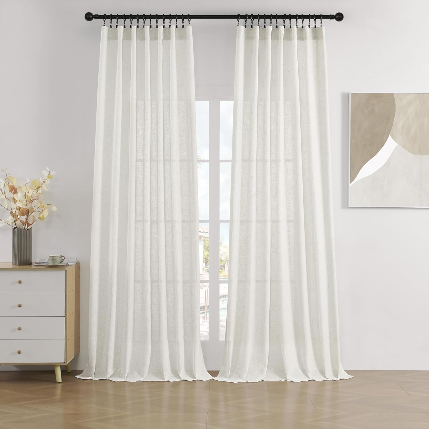 Two beige semi-sheer linen curtains with rod pockets hang on a black rod, filtering light into a room with a dresser and art.