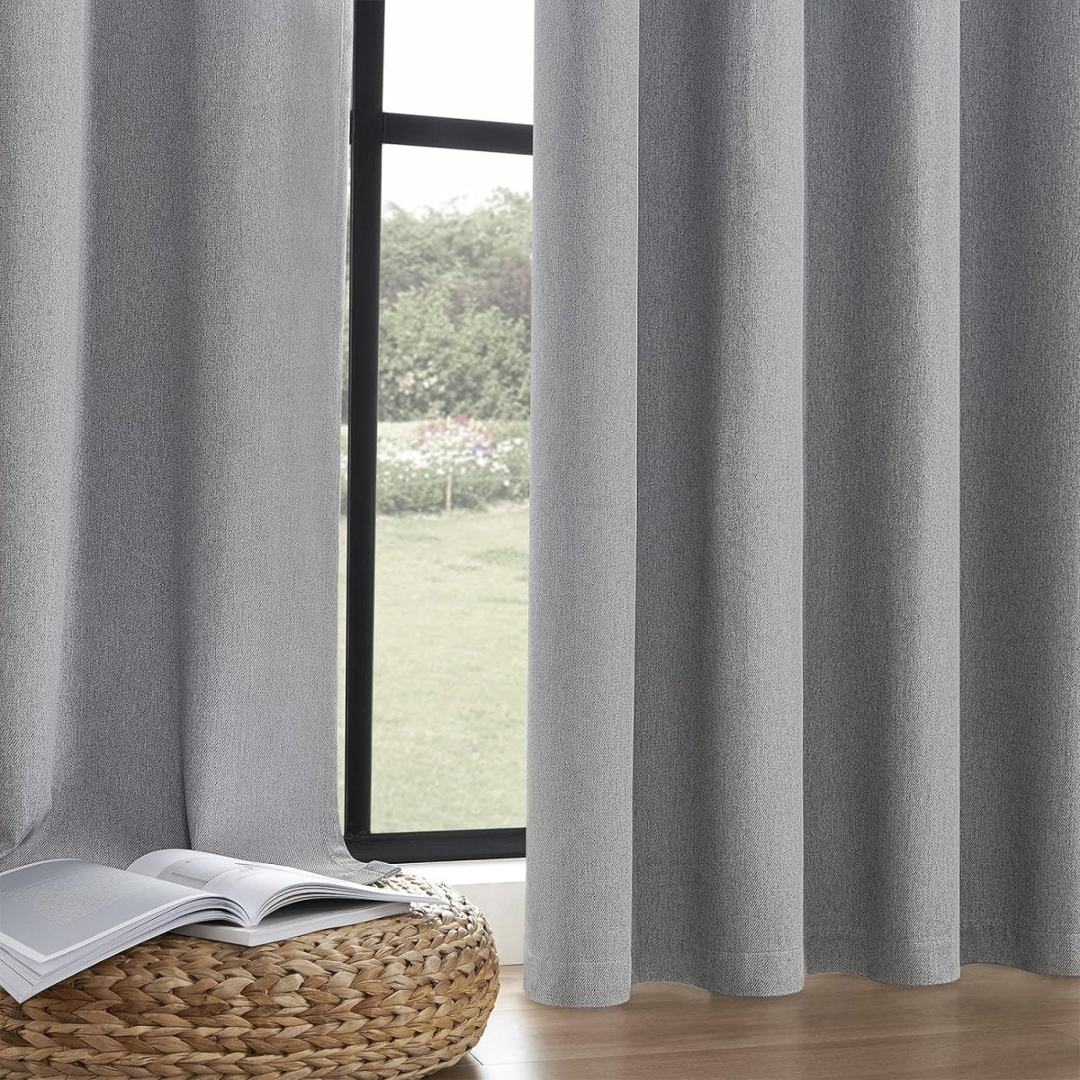 Two light gray Joydeco faux linen blackout curtain panels with grommets hang by a window, next to a stool with books.