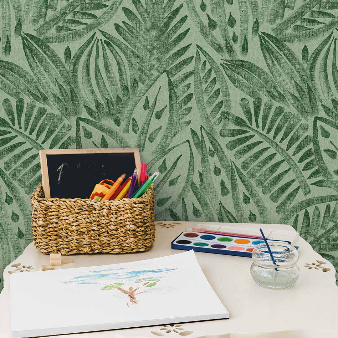 Tempaper's green grove Canvas Palm peel and stick wallpaper shown behind a kids play table with paint and drawings.