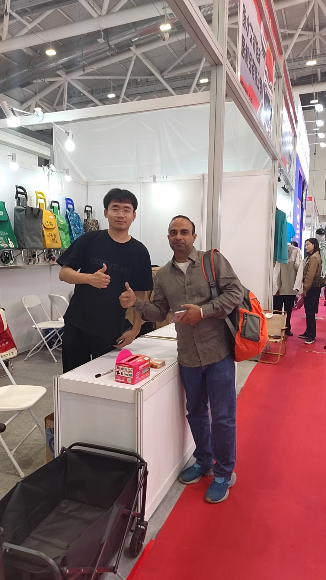 Chinese exhibitors posing for a group photo with foreign visitors at a luggage cart trade show booth, with folding trolleys and shopping carts displayed in the background.