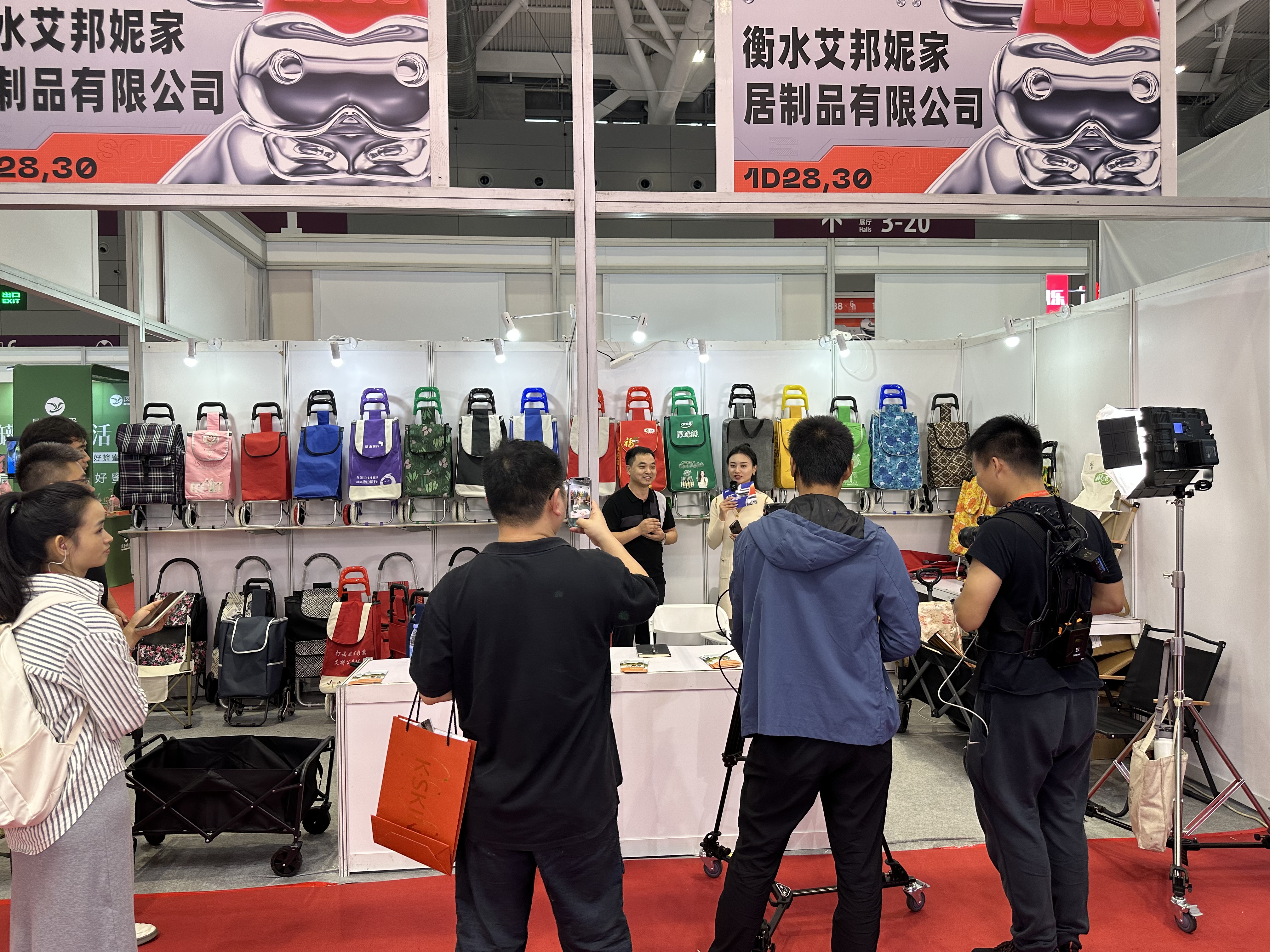 {"default":"A presenter live-streaming from a trade show booth, introducing and demonstrating a folding luggage cart to online viewers using a smartphone on a tripod."}
