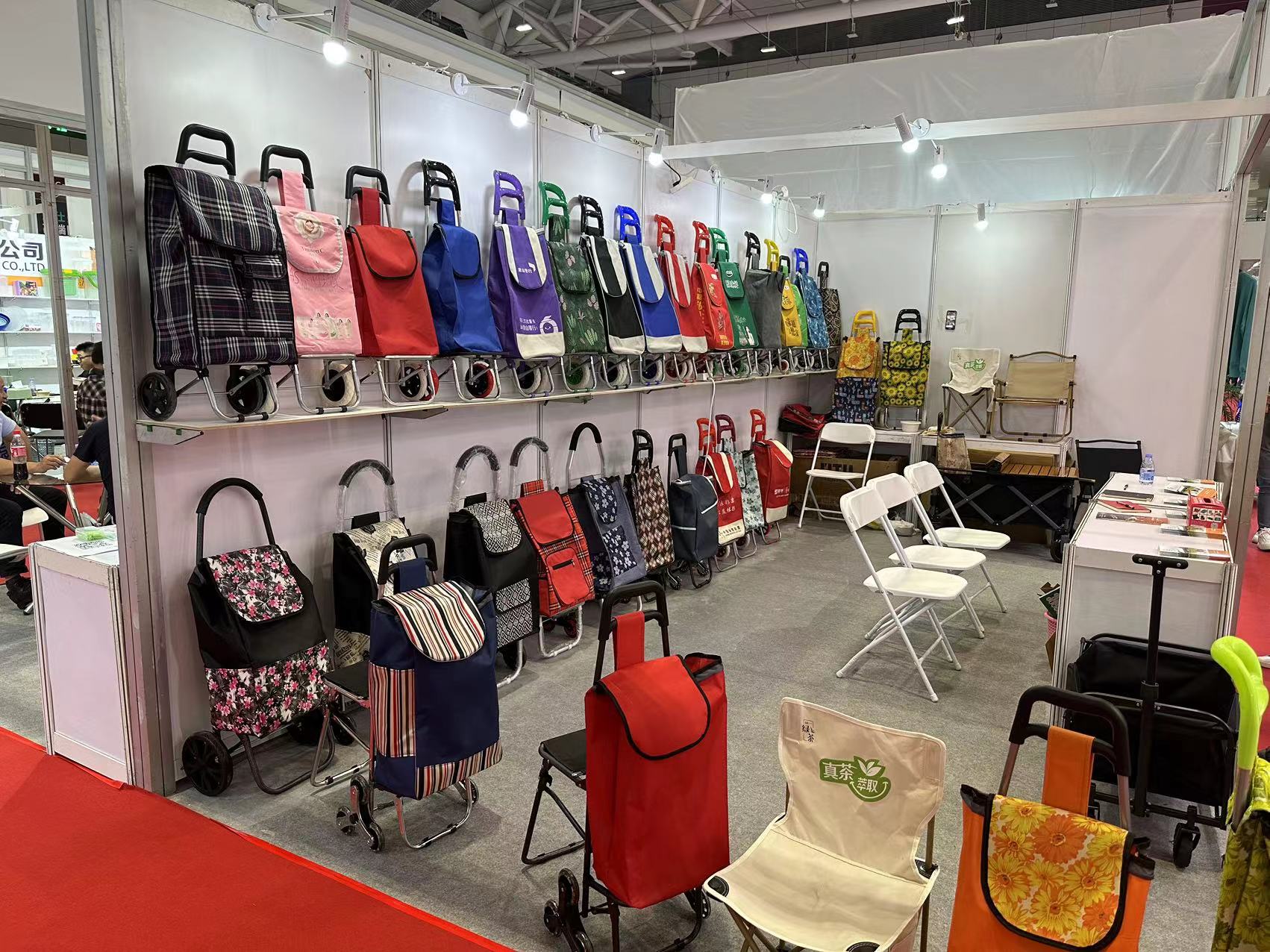 A variety of folding luggage carts and shopping trolleys are neatly arranged on shelves at a trade show exhibition booth, with different colors and sizes on display.