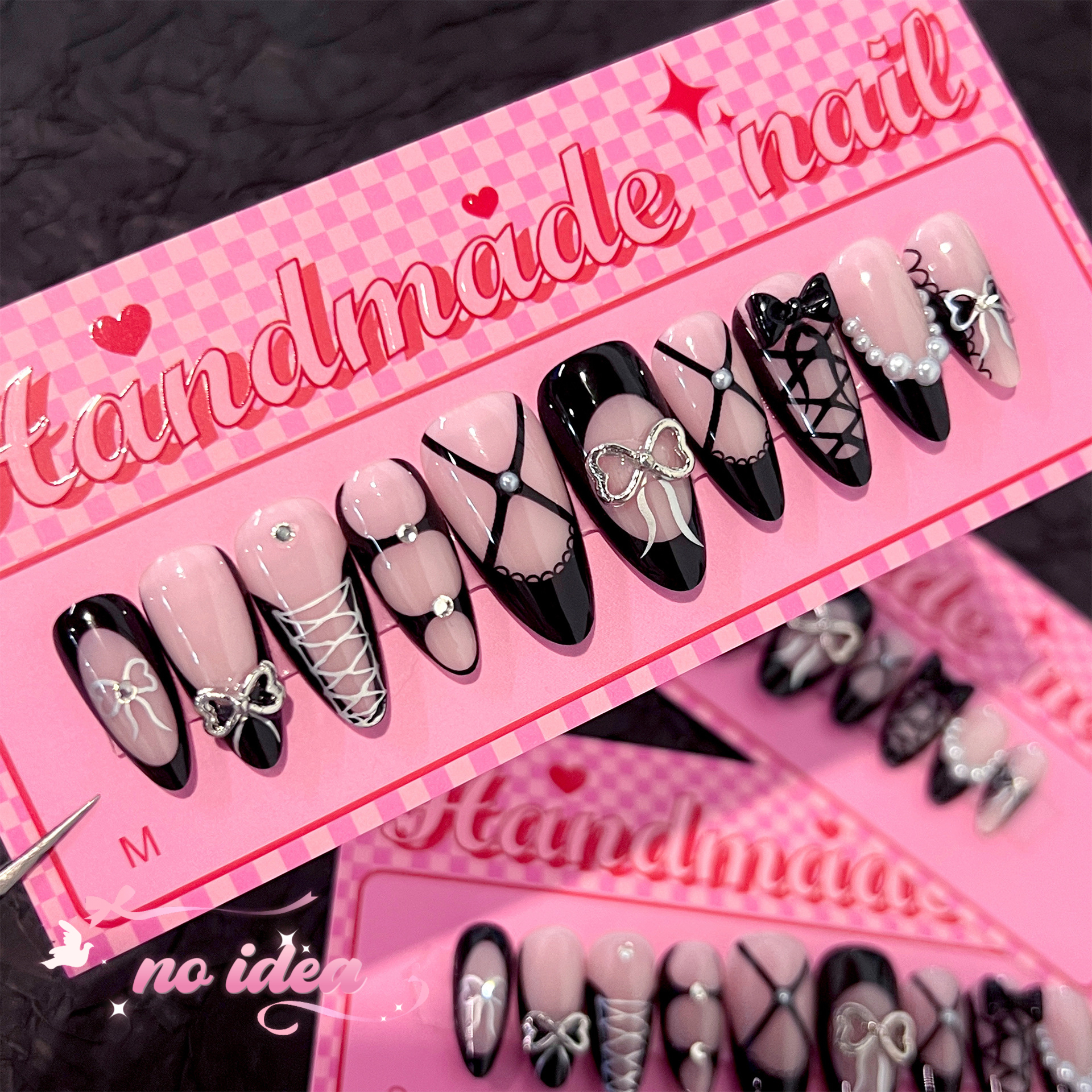 Long almond fake nails in black metallic with a large, reflective 3D silver bow on the ring finger, displayed with glue and tools.