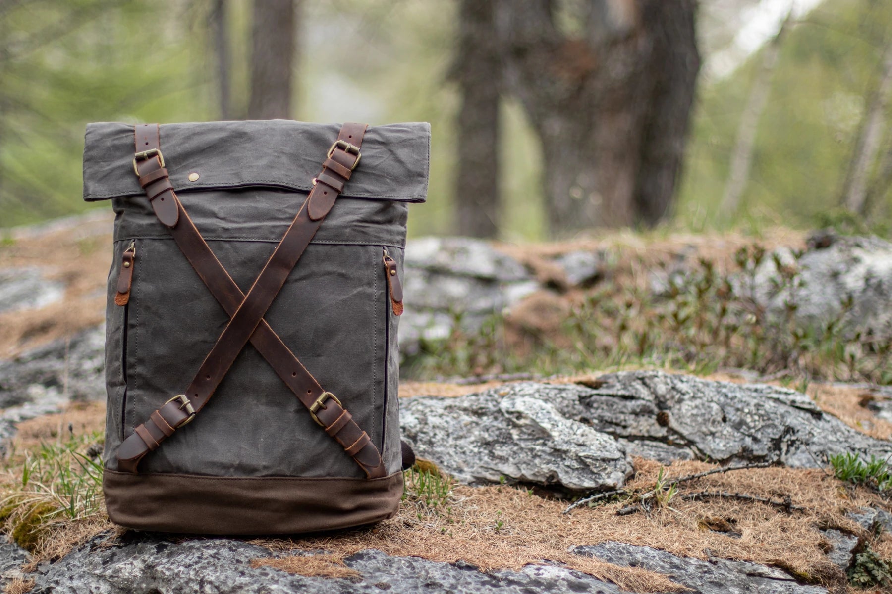 Dark grey Oslo waxed canvas rucksack standing on a moss-covered alpine rock in a pine forest. criss-cross leather strap closure and brown canvas base