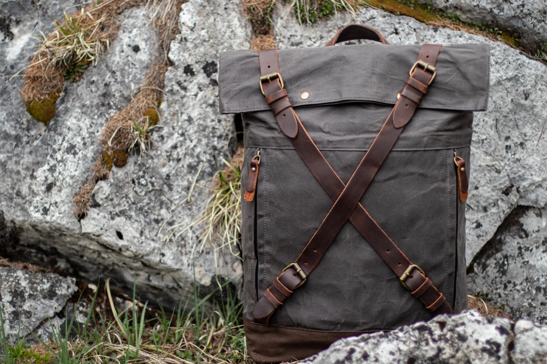Dark grey Oslo waxed canvas rucksack resting on alpine granite rocks. criss-cross leather strap closures and brass roller buckles catching daylight