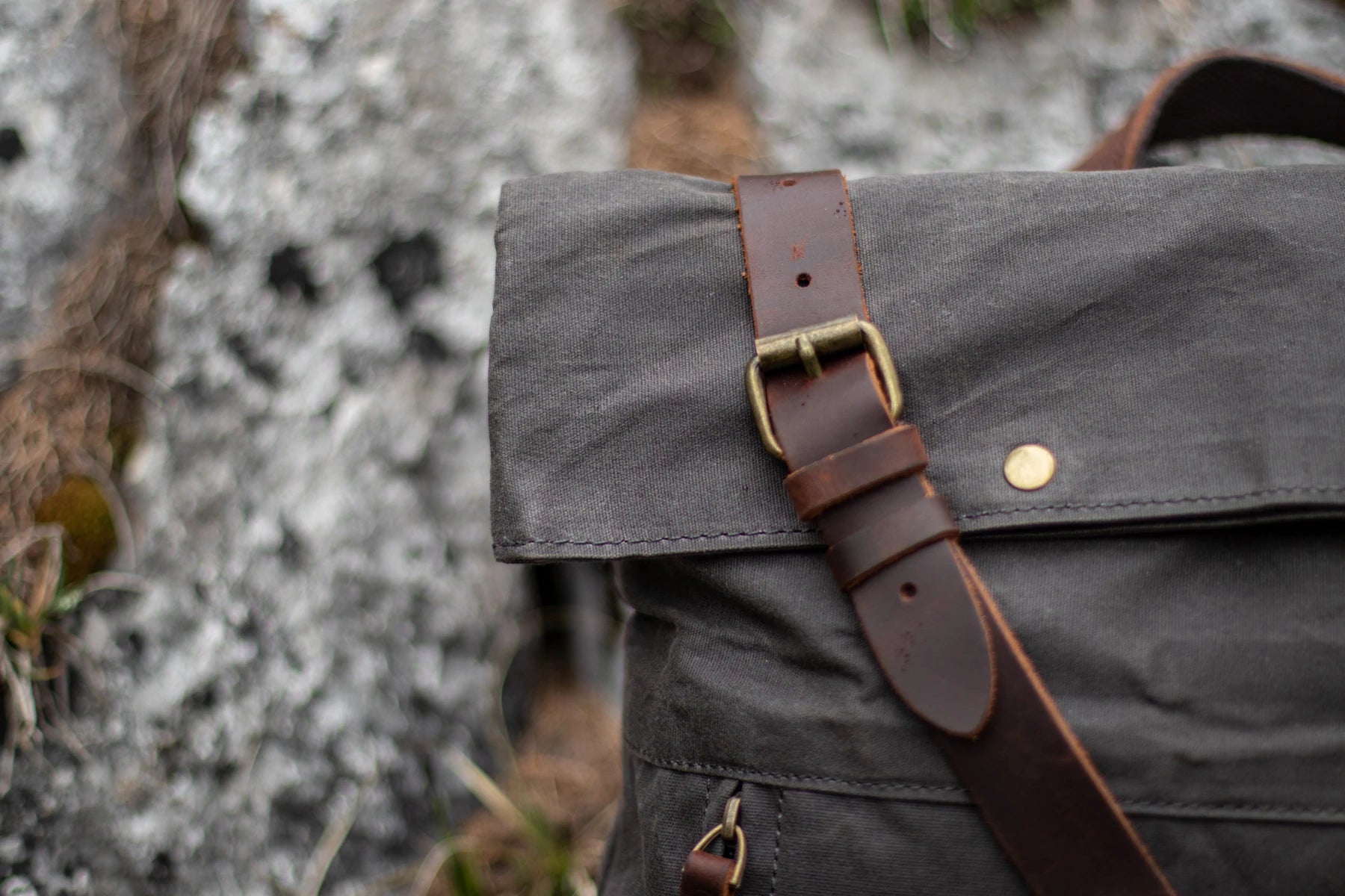 Macro of the Oslo top flap with full-grain leather strap. antiqued brass roller buckle and brass rivet. against a soft alpine rock backdrop