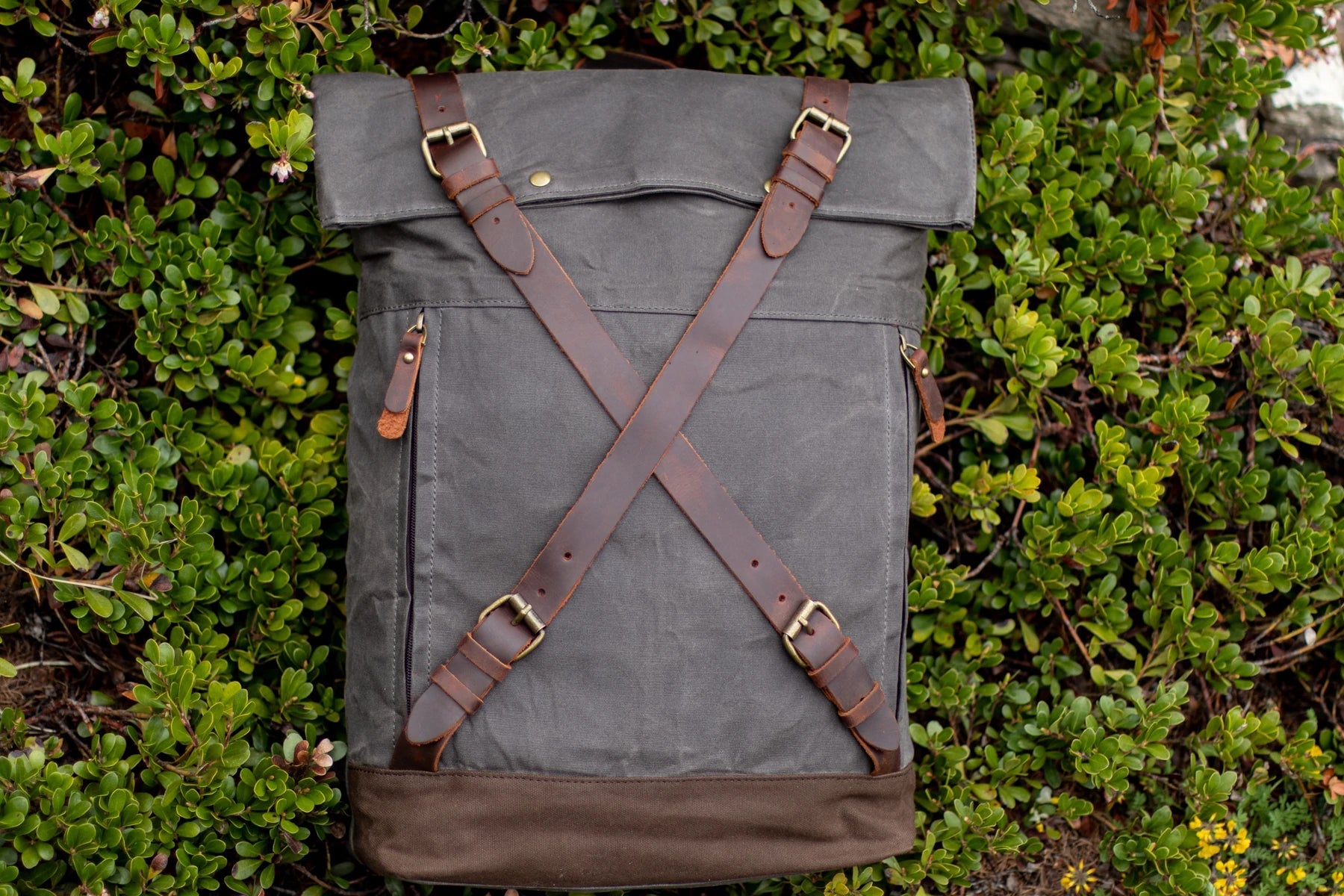 Top-down flat-lay of the dark grey Oslo canvas rucksack on bilberry foliage. criss-cross full-grain leather straps with antiqued brass buckles and side zip access