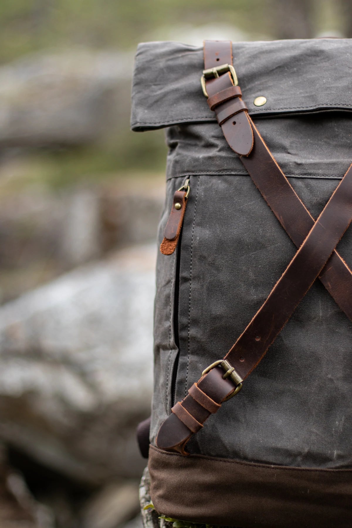Vertical detail of the criss-cross full-grain leather straps on the dark grey Oslo waxed canvas rucksack. brass buckles and side zip pocket pull visible