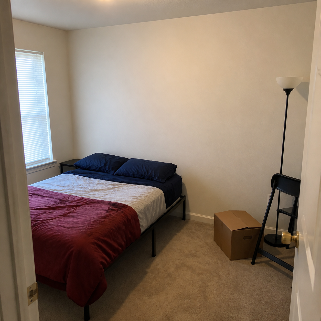 AI-generated illustration of a fictional bedroom with a metal bed frame, folding chair, floor lamp, boxes and a blank wall above the bed.