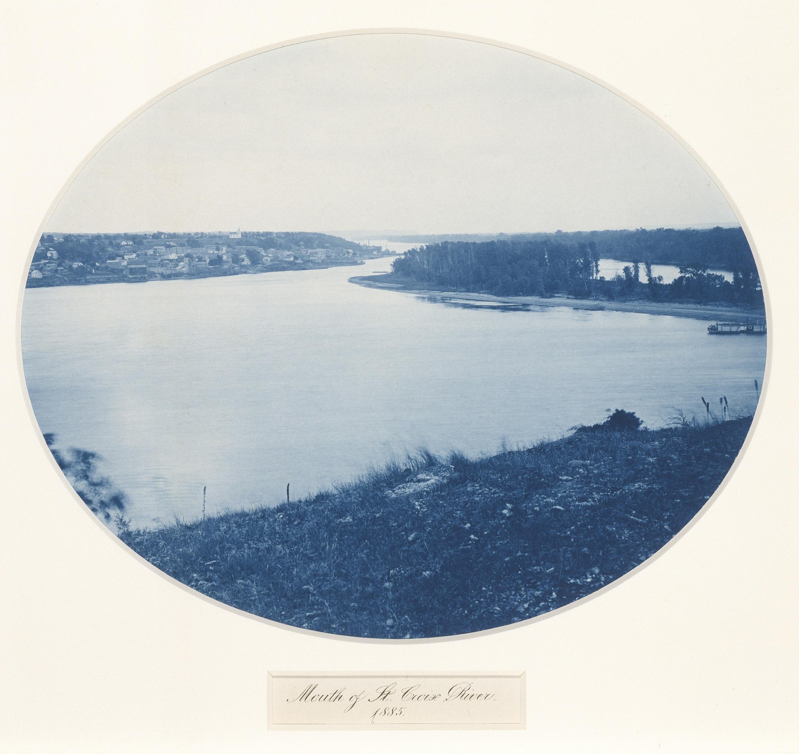 An oval blue photograph shows a broad river, buildings on the far left bank, a wooded spit at right and a dark grassy bank in the foreground.