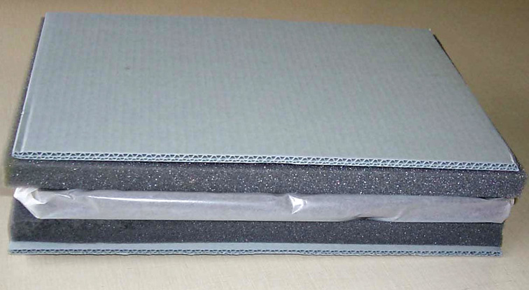 A wrapped rectangular frame sits between dark foam sheets and corrugated boards above and below.
