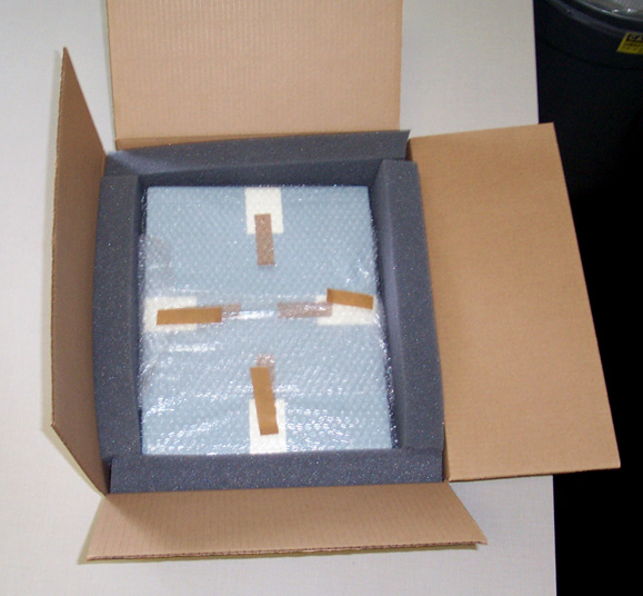A bubble-wrapped rectangular package sits inside an open cardboard box with thick dark foam around its sides.