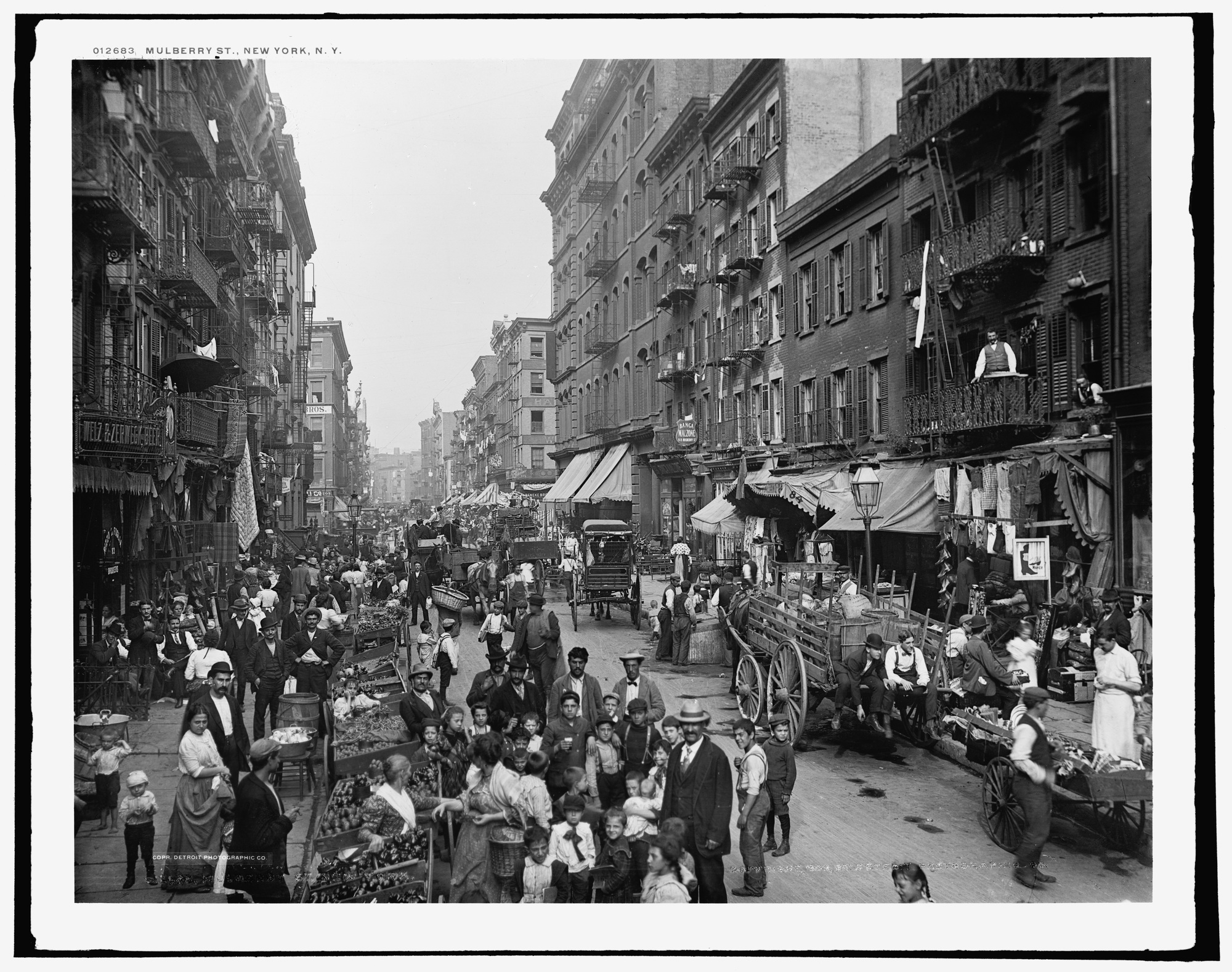 A black-and-white photograph looks down Mulberry Street past market carts and pedestrians, with rows of buildings, shopfronts and fire escapes receding into the distance.