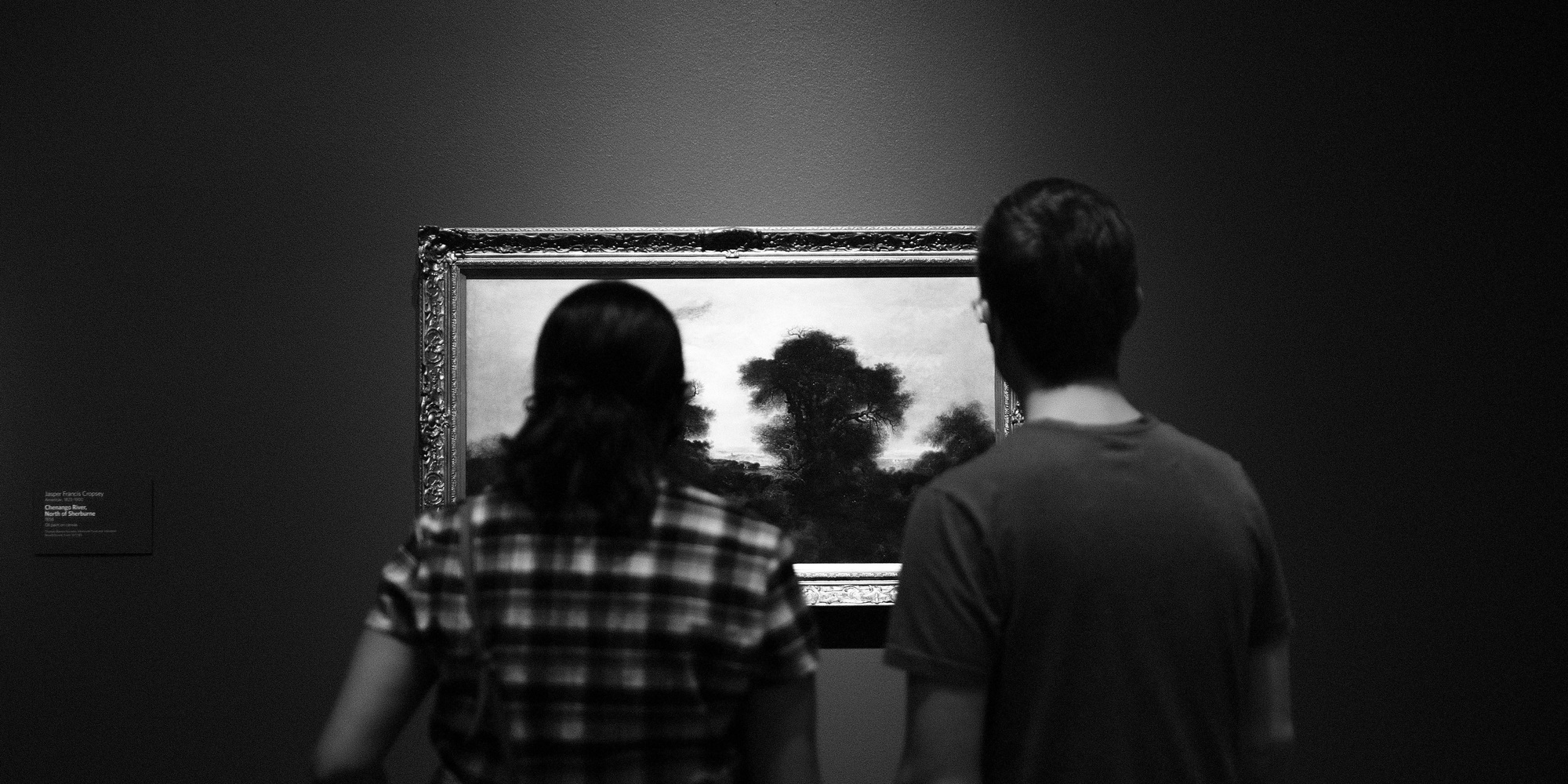 Two gallery visitors stand before a brightly pictured framed landscape on a dark wall in a black-and-white photograph.
