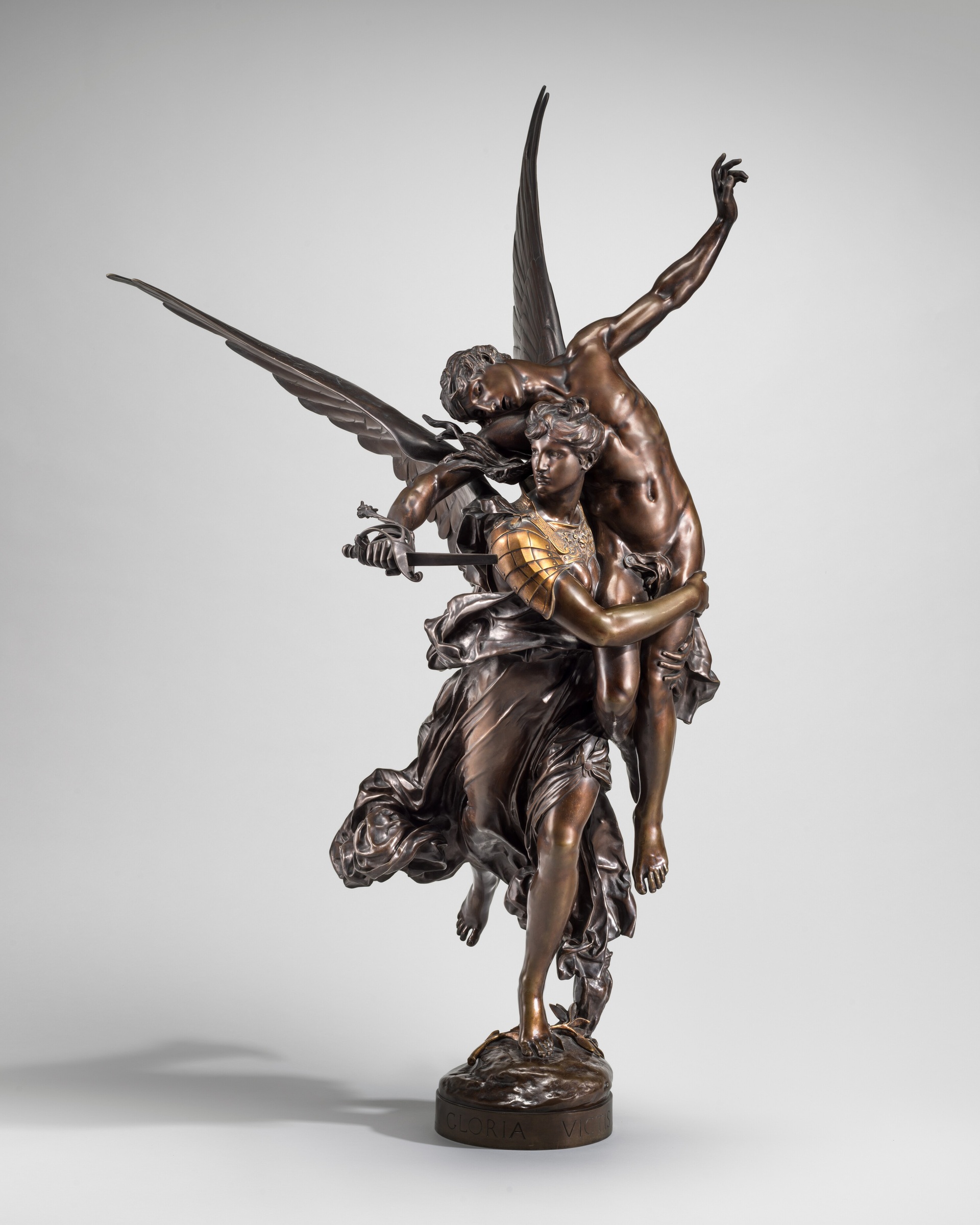 Bronze winged figure carrying a fallen man with a broken sword against a plain grey background.