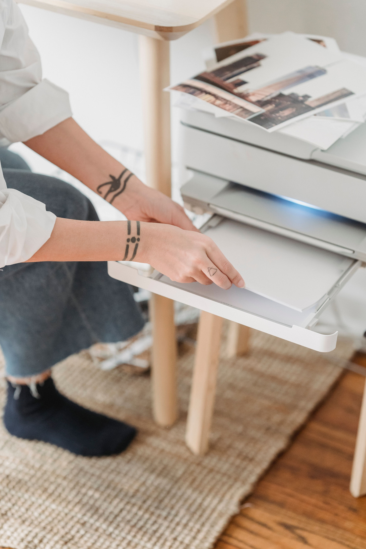 Hands place a stack of white paper into an open printer tray, with printed city photographs resting on top of the printer.