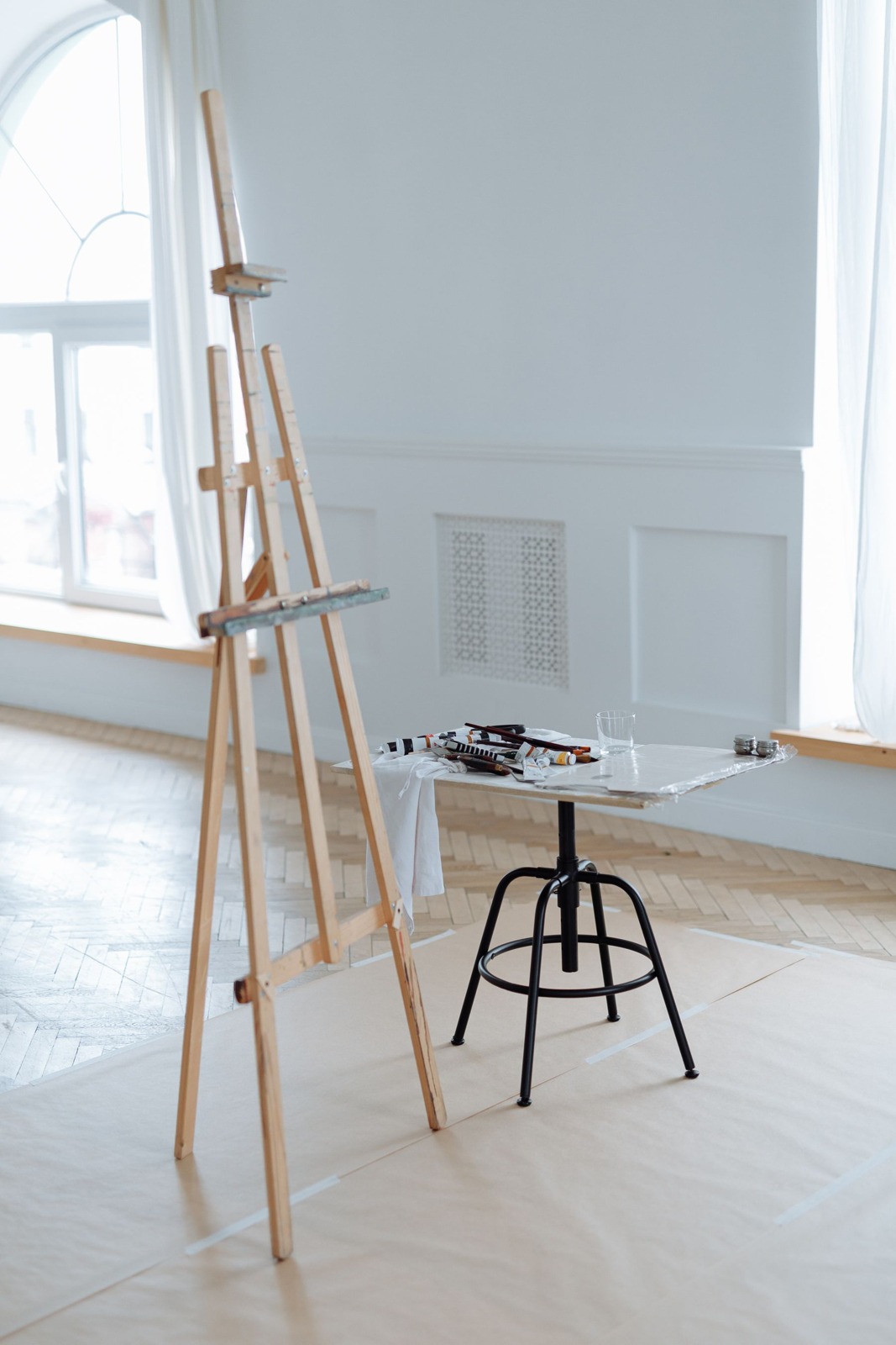 An empty wooden easel and painting supplies beside large studio windows.