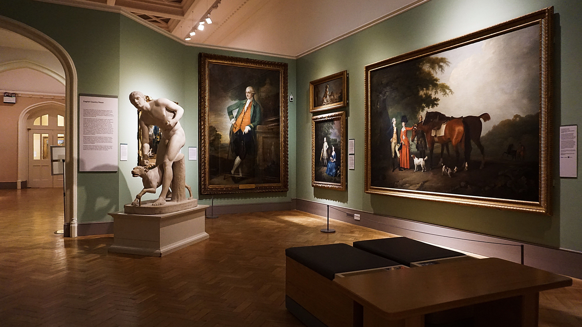 An empty art gallery with green walls, framed classical paintings, a white marble sculpture and wooden benches.