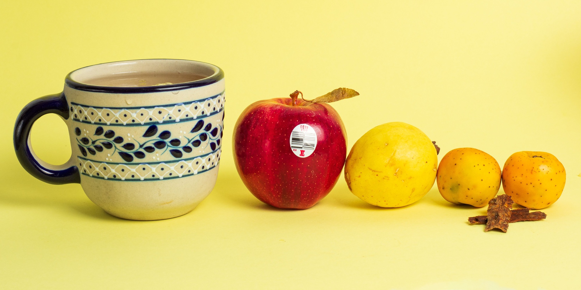 A patterned ceramic mug, red apple and yellow fruits sit in a row against a yellow backdrop.