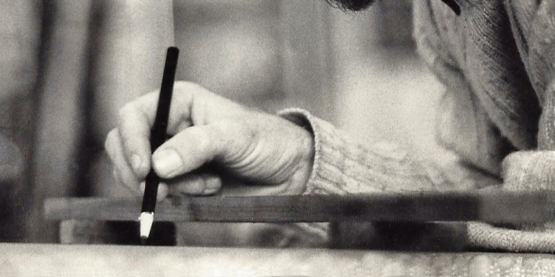 Black-and-white detail of a hand holding a lithographic pencil over a stone, with the artist’s arm and beard at the edge.