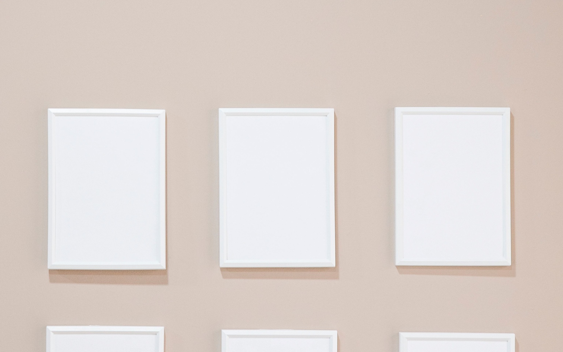 Three empty white frames on a warm gray wall, with the top edges of a lower row visible.