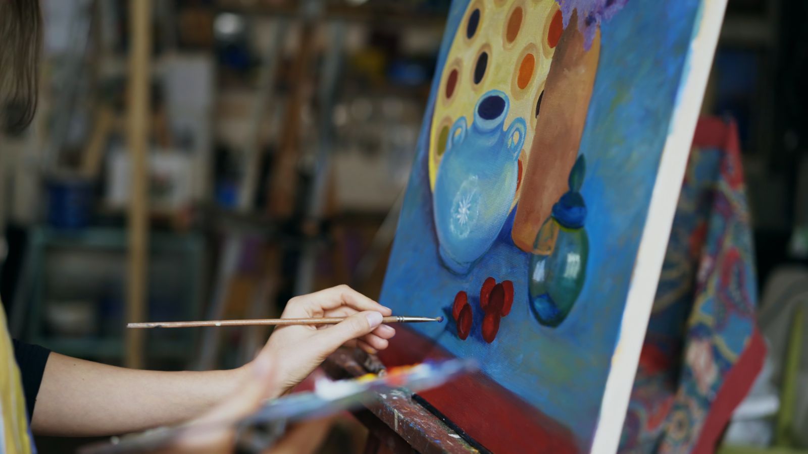A hand holding a fine paintbrush beside a colorful still-life painting on an easel.