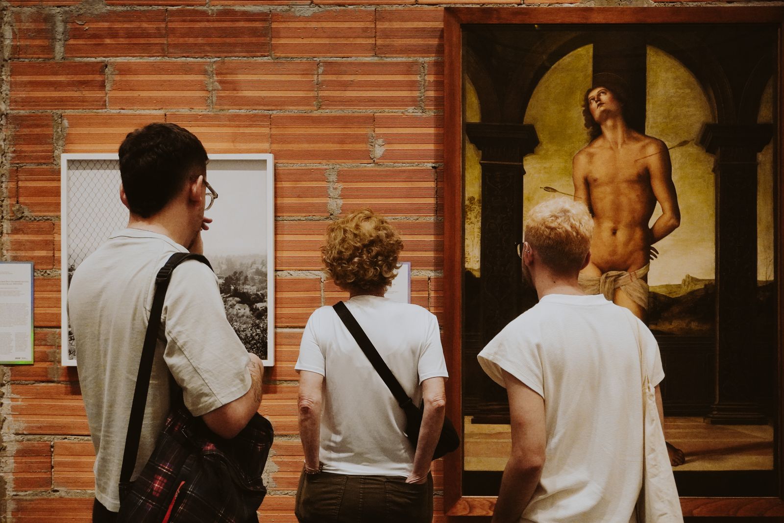 Three gallery visitors seen from behind, looking at a framed painting on a brick wall.