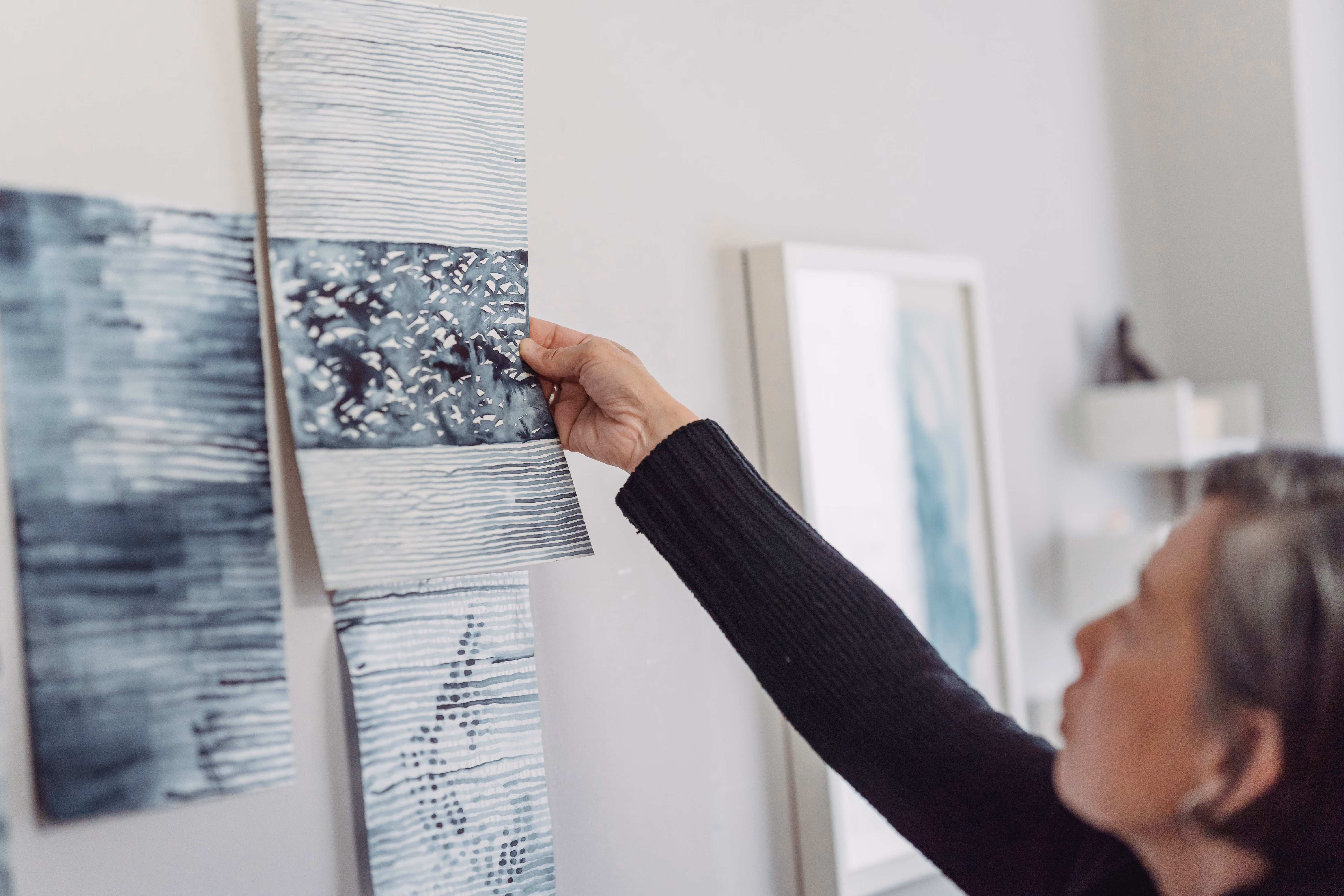 A woman compares abstract paintings on a wall, pausing to examine individual image studies.
