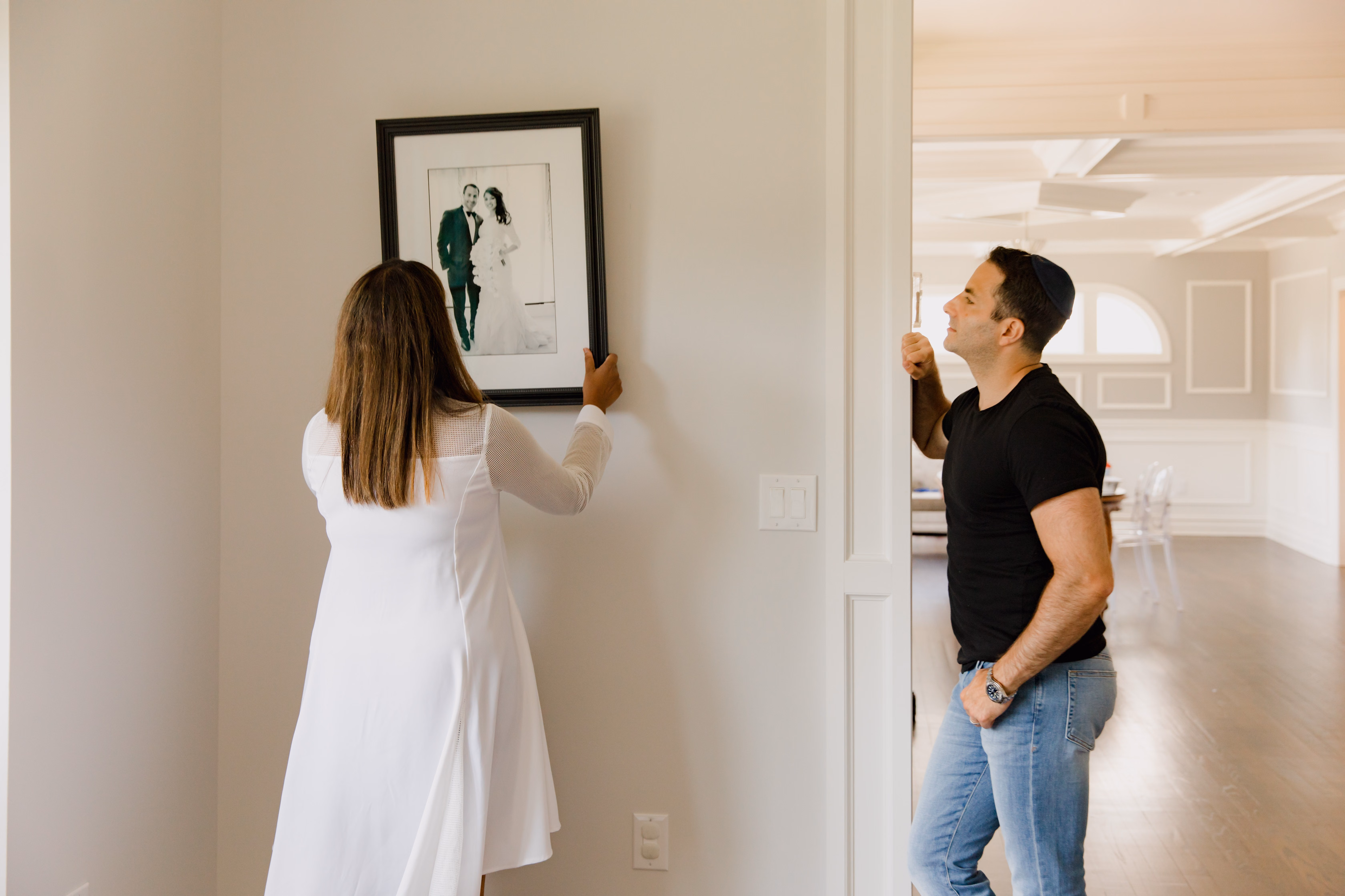 A couple decorating their home by holding a framed photograph near a wall.