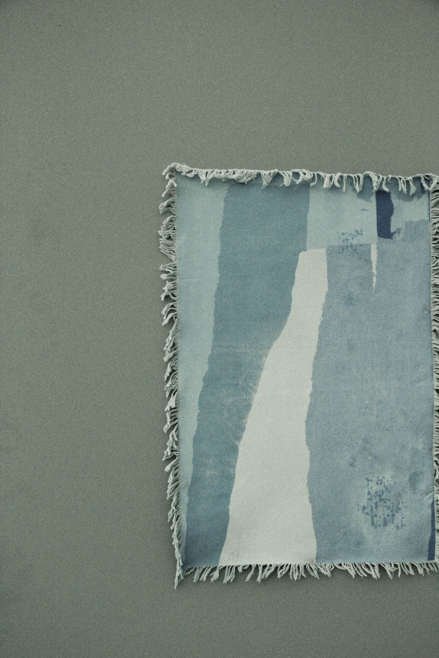 Blue and pale abstract textile with a fringed edge hanging on a muted wall.