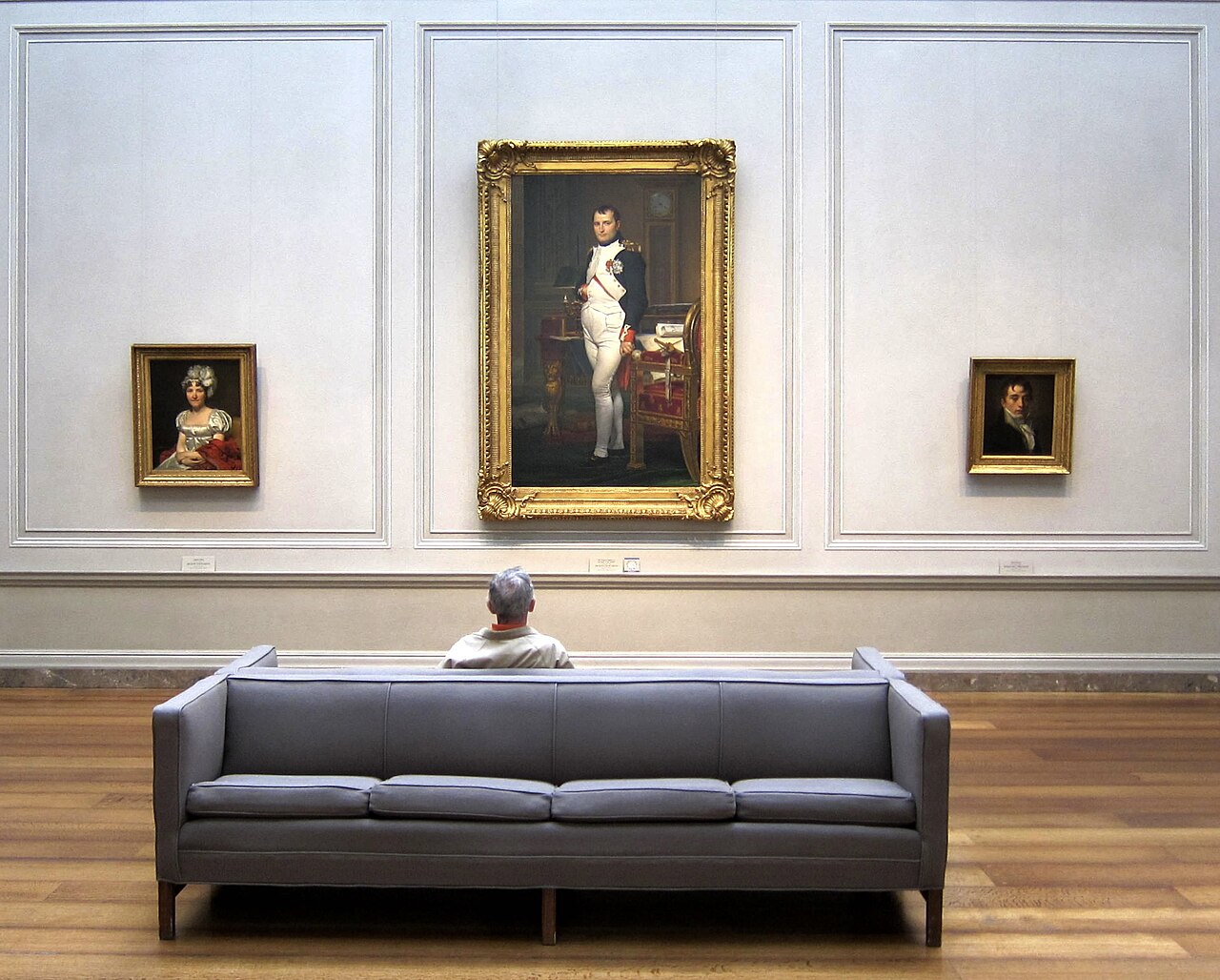 A visitor sits behind a gray sofa facing a large gold-framed full-length portrait of Napoleon, with two smaller framed portraits on the pale wall beside it.
