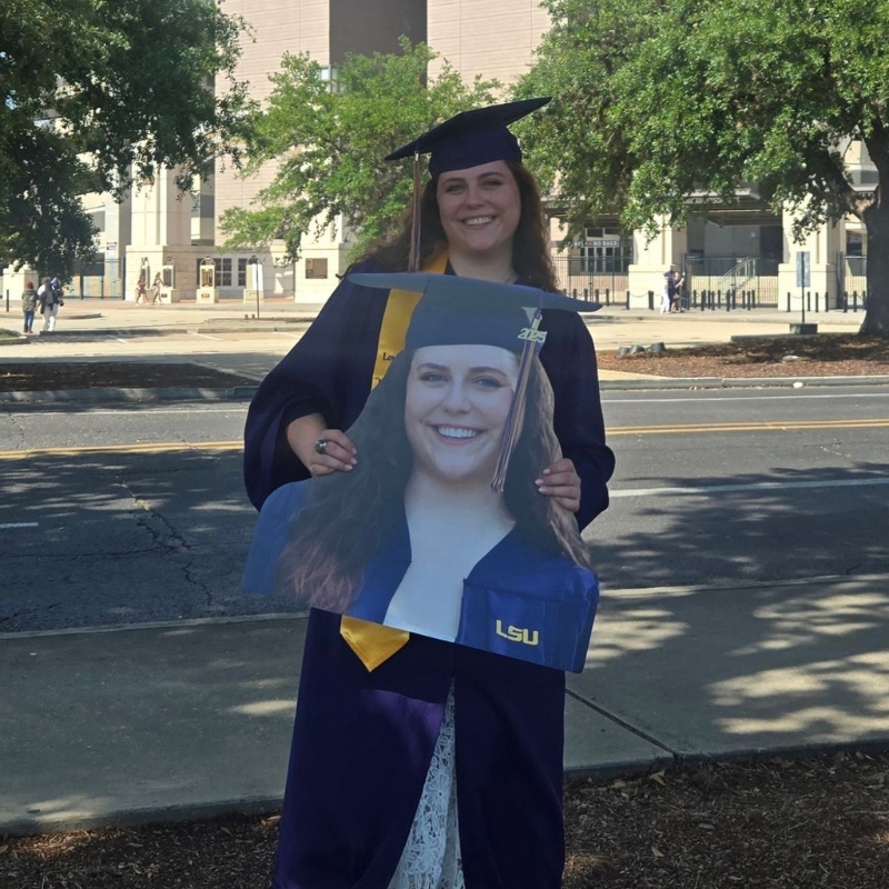 Graduate in cap and gown holding her own big head cutout on campus