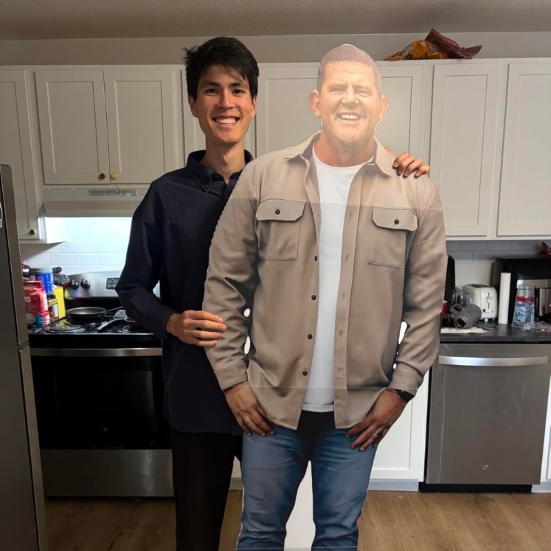 Man smiling and posing with his friend's life-size cutout in home kitchen