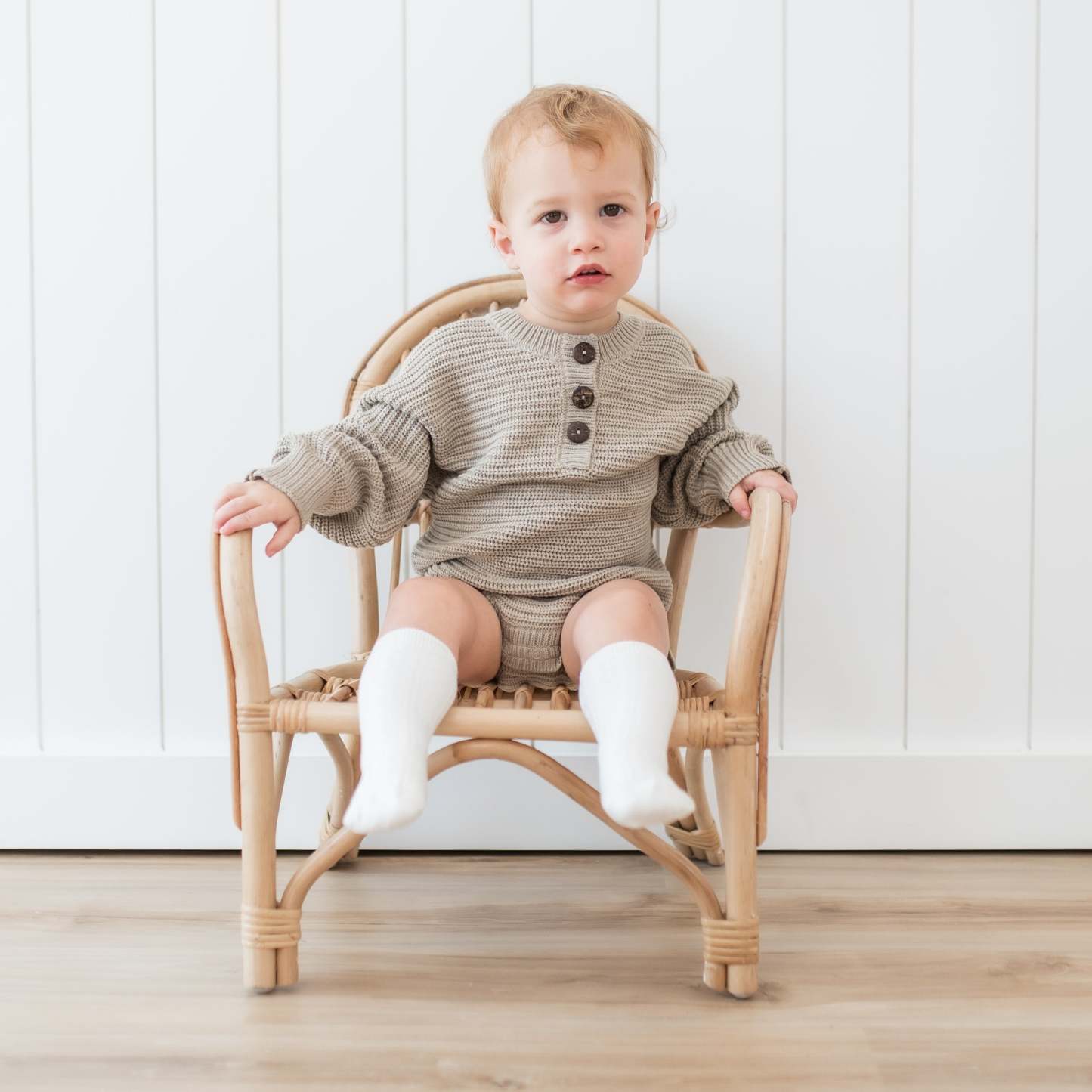 Toddler sitting in a wicker chair wearing the Chunky Knit Romper in Almond and Cloud Knee high socks