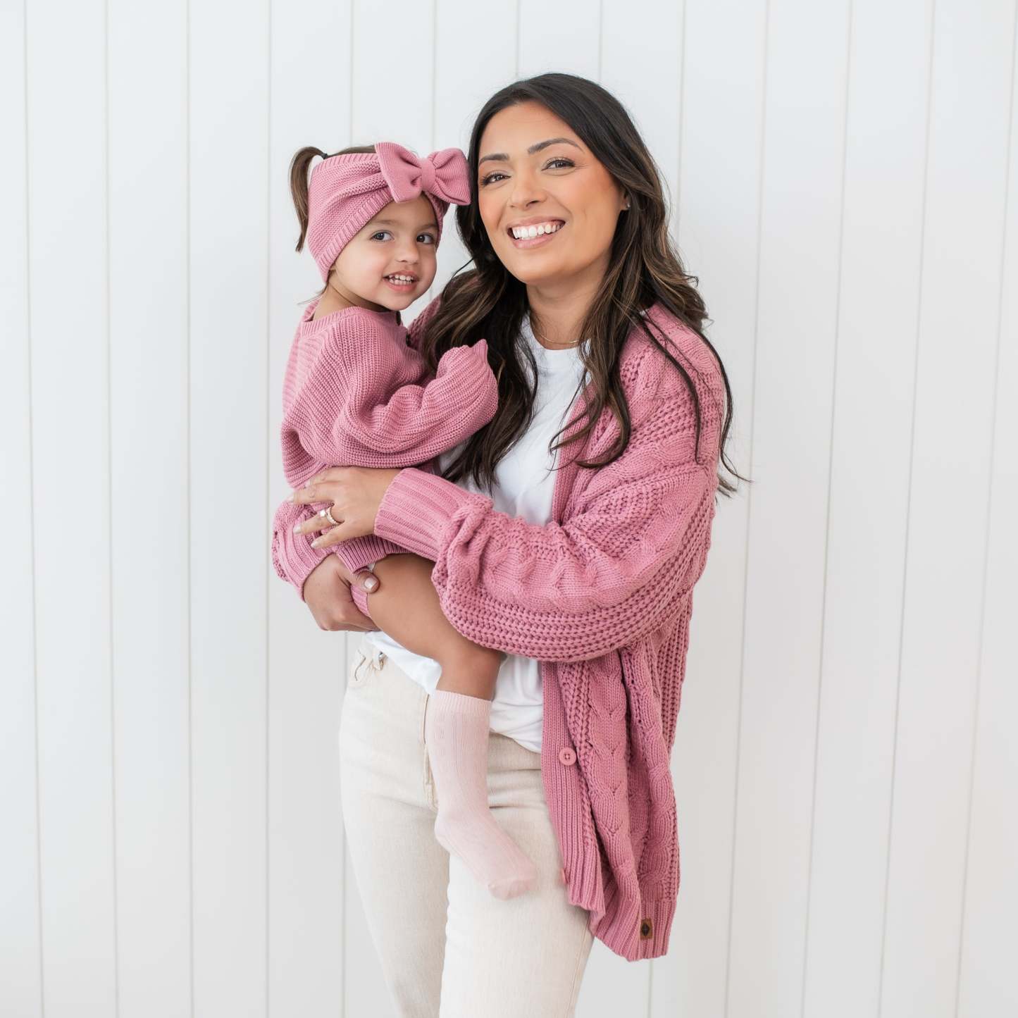 Smiling mother wearing the Cable Knit Cardigan in Apple blossom holding her daughter who is wearing the Chunky Knit Romper in Apple Blossom and matching chunky knit bow headband