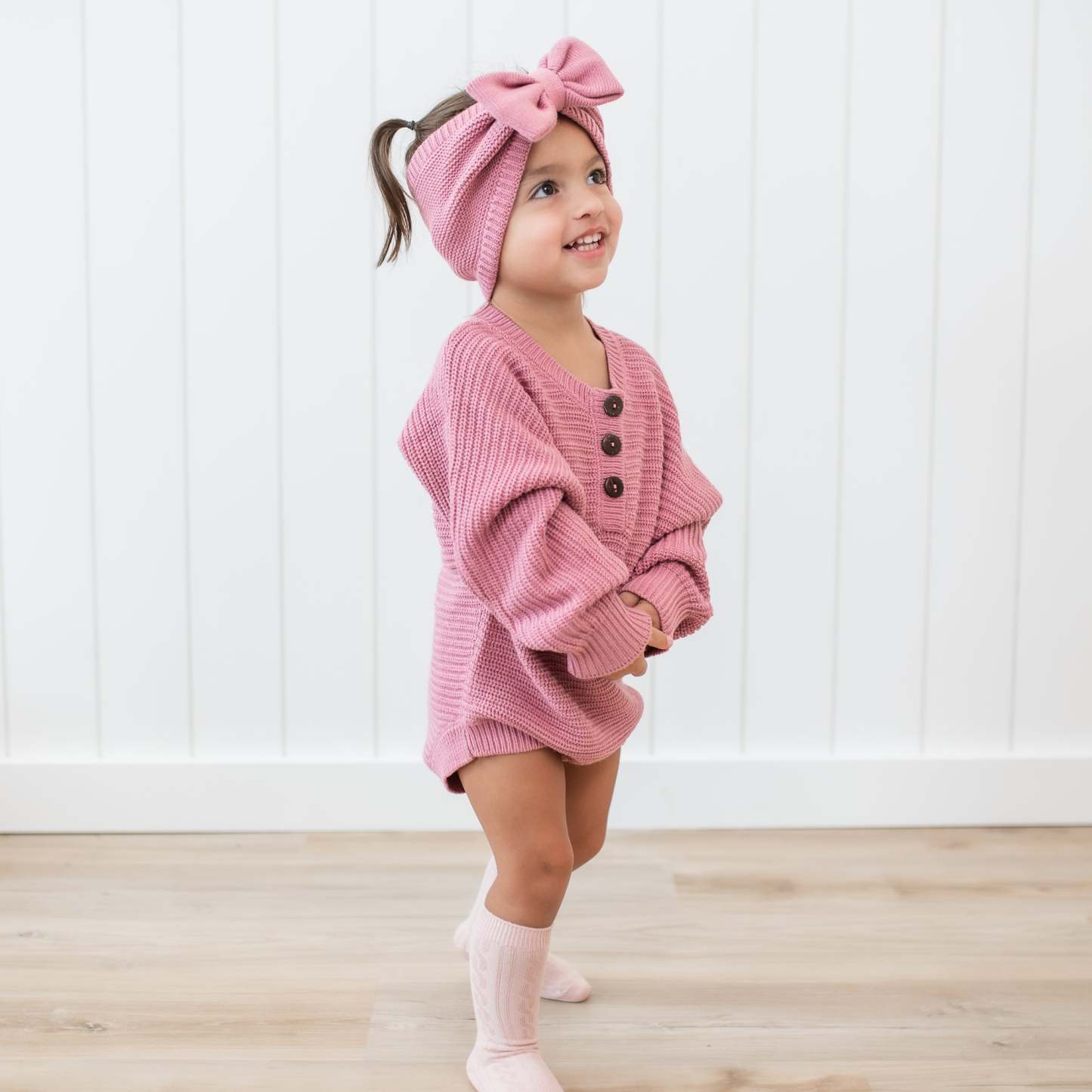 Smiling toddler wearing the Chunky Knit Romper in Apple Blossom with matching chunky knit bow headband