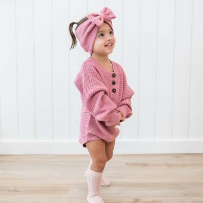 Smiling toddler wearing the Chunky Knit Romper in Apple Blossom with matching chunky knit bow headband