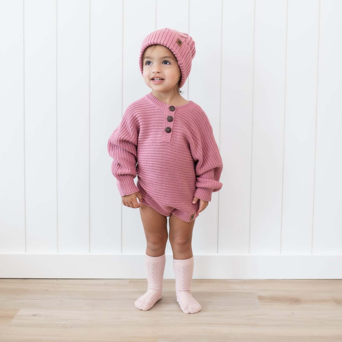 Toddler wearing the Chunky Knit Romper in Apple Blossom with matching cable knit pom beanie standing in front of a white paneled wall