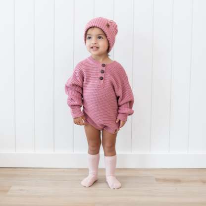 Toddler wearing the Chunky Knit Romper in Apple Blossom with matching cable knit pom beanie standing in front of a white paneled wall