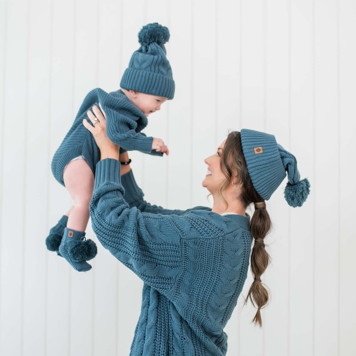 Mother holding toddler in the air both wearing chunky knit items in the color Atlantic