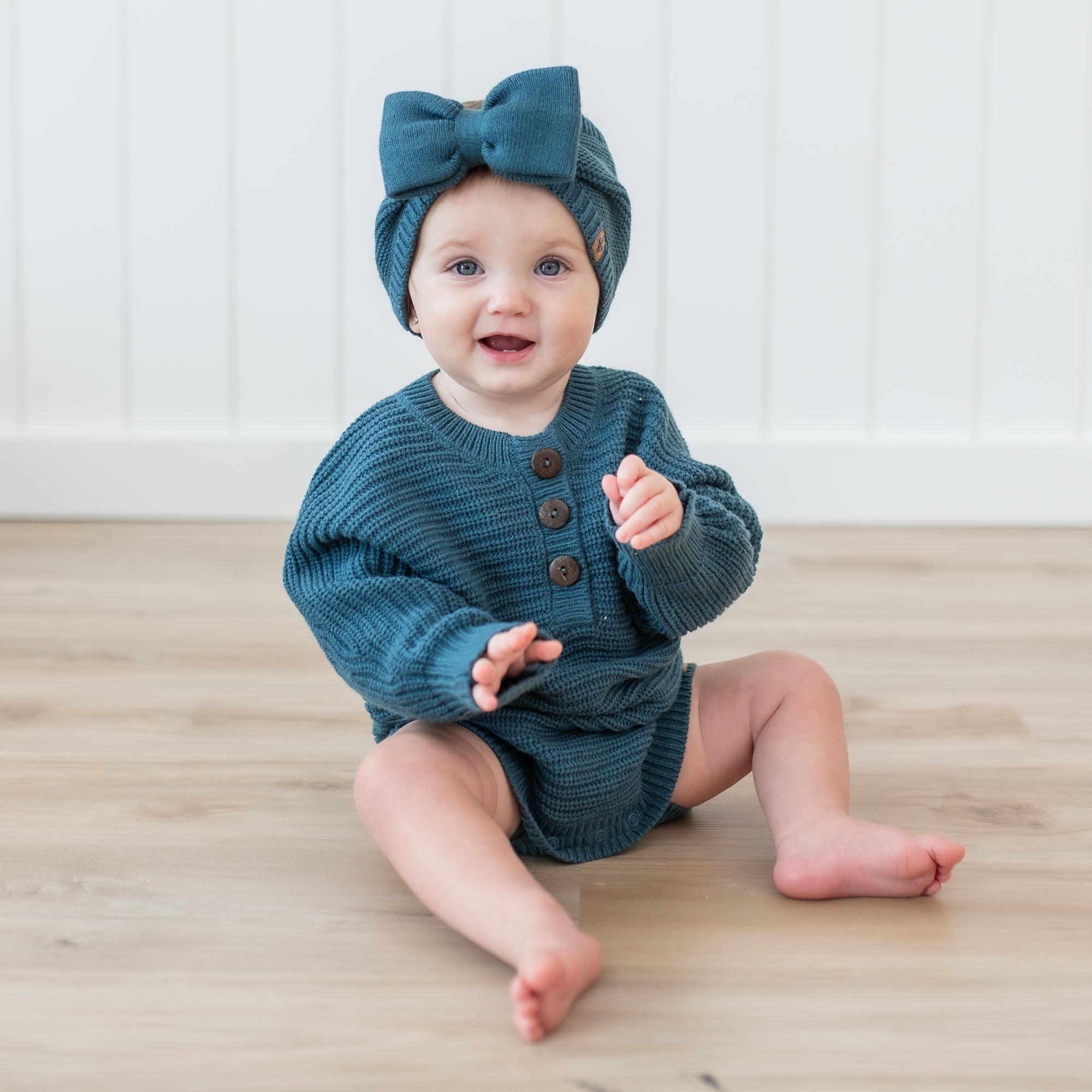 Toddler sitting on the floor wearing the Chunky Knit Romper in Atlantic paired with a Chunky knit bow headband