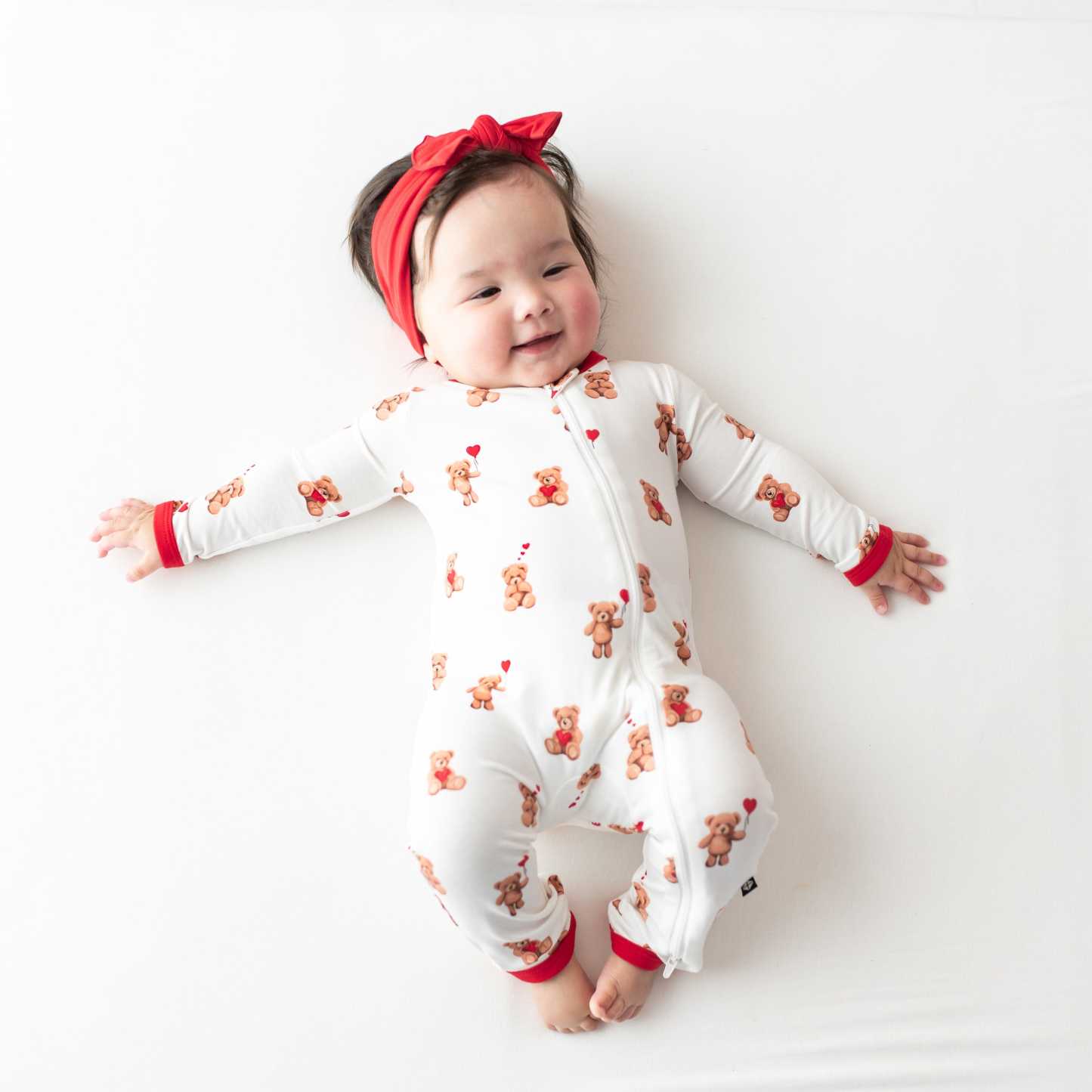 Smiling infant lying down wearing the  soft and breathable Zippered Romper in Bear Hearts paired with a Cardinal knotted bow headband