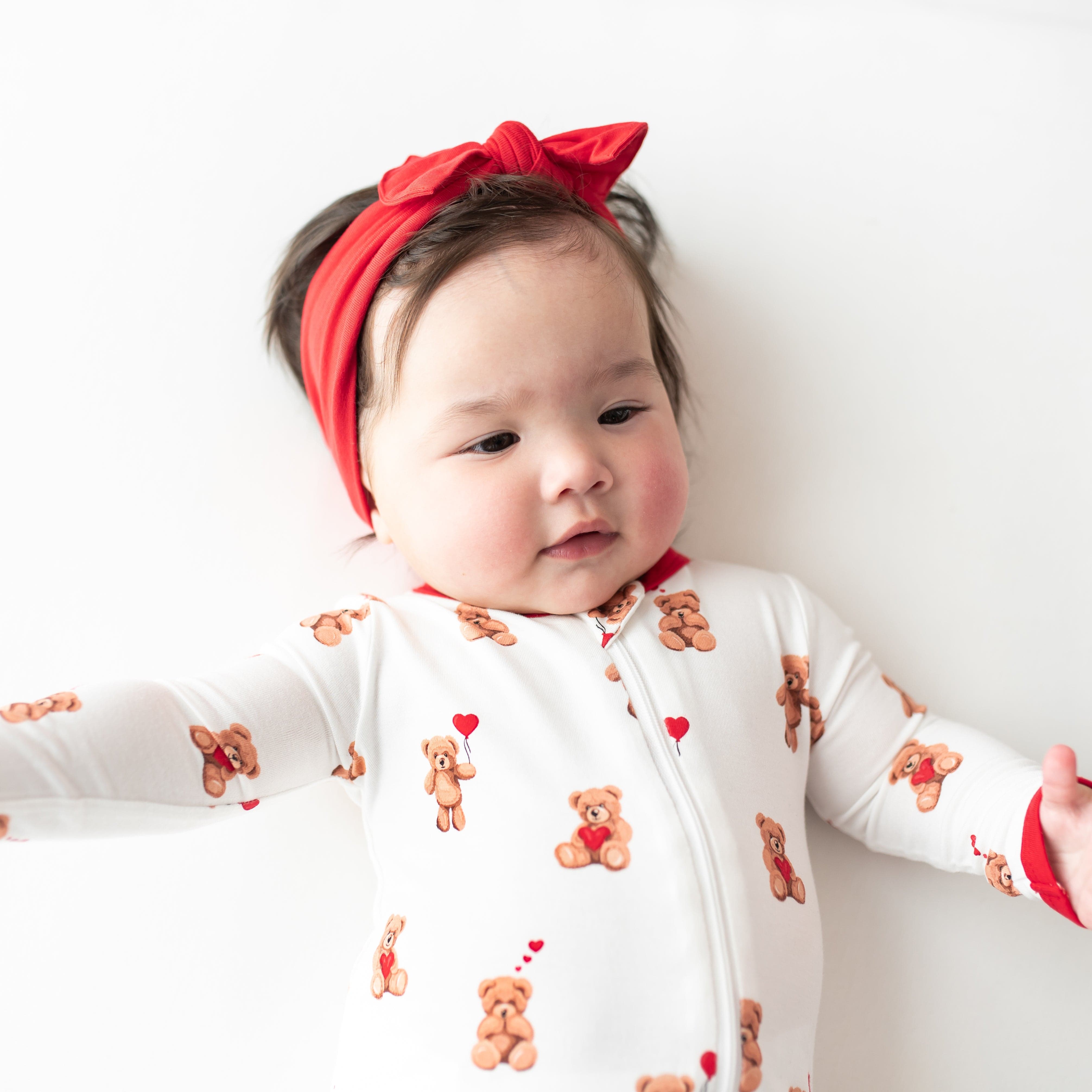 Close up of infant wearing the Zippered Romper in Bear Hearts with Cardinal knotted bow headband